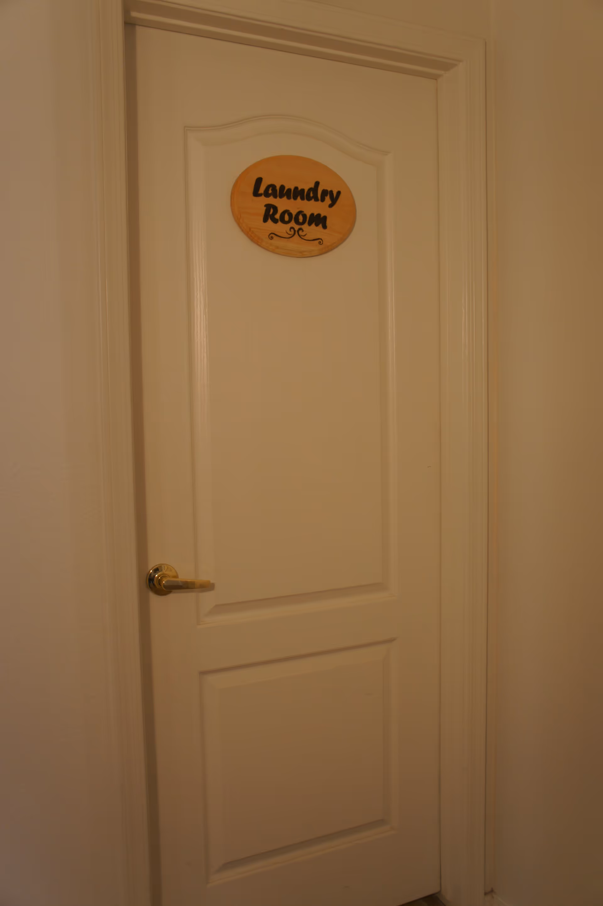 A closed white door with a gold-colored handle and a wooden oval sign that reads 'Laundry Room' mounted on the upper center of the door.