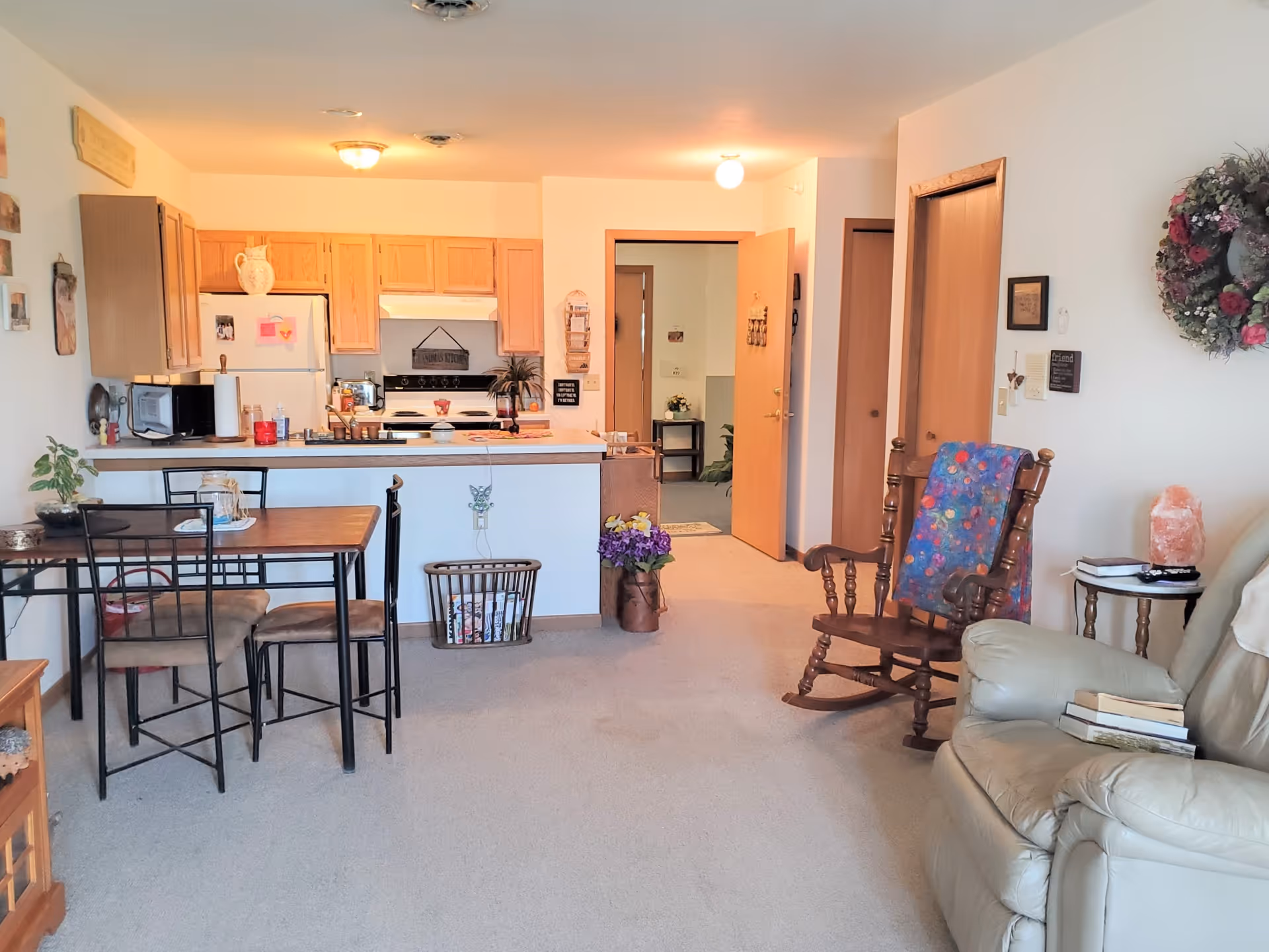 Interior view of a senior living apartment showing a small dining area with a table and four chairs, a kitchen with wooden cabinets, a white refrigerator, and a stove. There is a living area with a beige recliner, a wooden rocking chair with a colorful blanket, a small side table with books and a lamp, and a floral wreath on the wall. The room is carpeted and has an open doorway leading to another room.