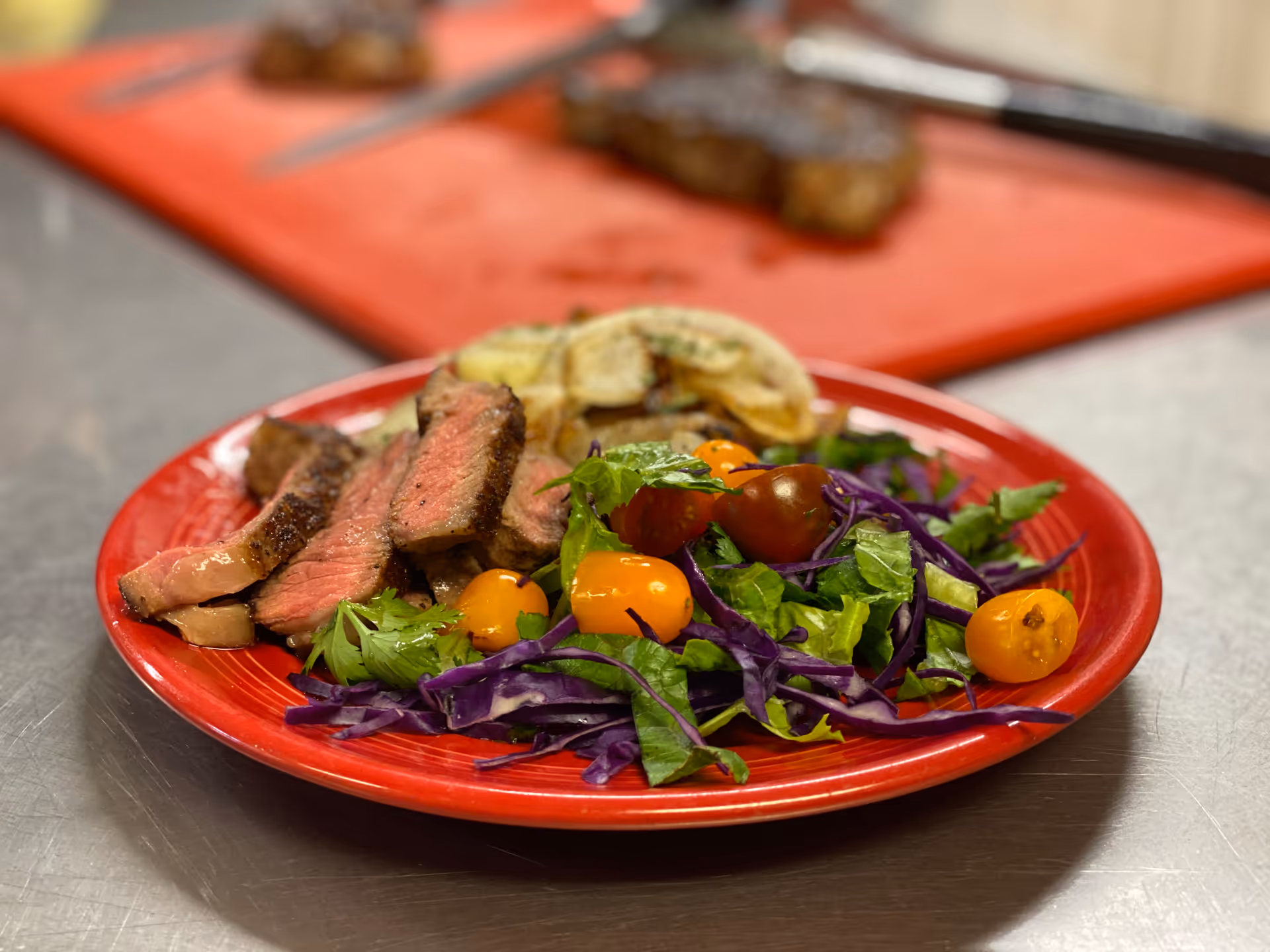 A red plate with slices of medium-rare steak, a fresh salad with cherry tomatoes, purple cabbage, and leafy greens, and some grilled potato slices in the background on a stainless steel surface.