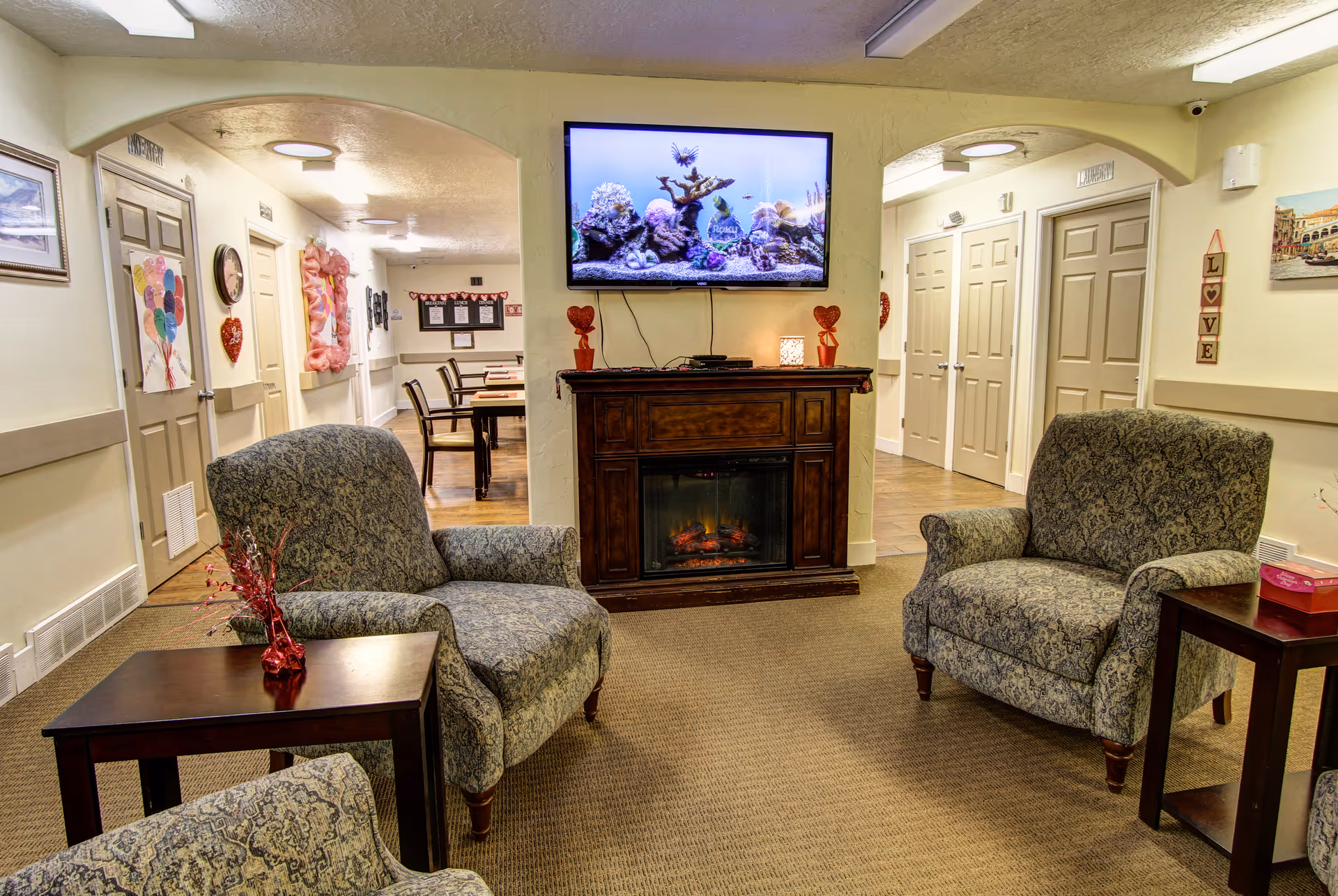 A cozy common area in a senior living facility with three patterned armchairs arranged around two small wooden side tables. A wooden electric fireplace with a TV mounted above it is centered on the far wall. The TV displays an aquarium scene. The room has beige walls and carpet, with doorways leading to other rooms and a hallway with tables and chairs visible in the background. Decorative items include red heart-shaped ornaments on the fireplace and wall art.