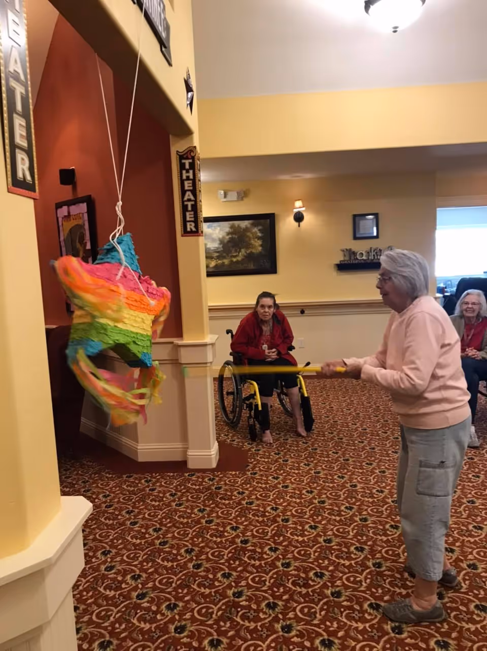 An elderly woman in a pink sweater and gray pants is hitting a colorful star-shaped piñata with a stick inside a room with patterned carpet. Another elderly woman in a red jacket sits in a wheelchair watching, and a third elderly woman is seated in the background smiling. The walls are painted yellow and decorated with framed pictures and theater signs.
