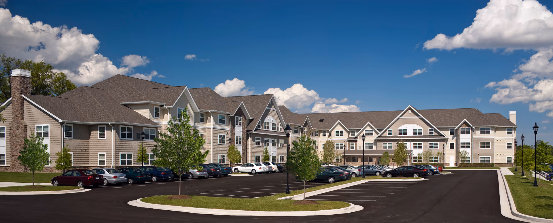 Exterior view of a large, multi-story senior living facility building with beige and brown siding, multiple windows, and a spacious parking lot with several cars parked. The sky is blue with scattered clouds, and there are small trees and lamp posts around the parking area.