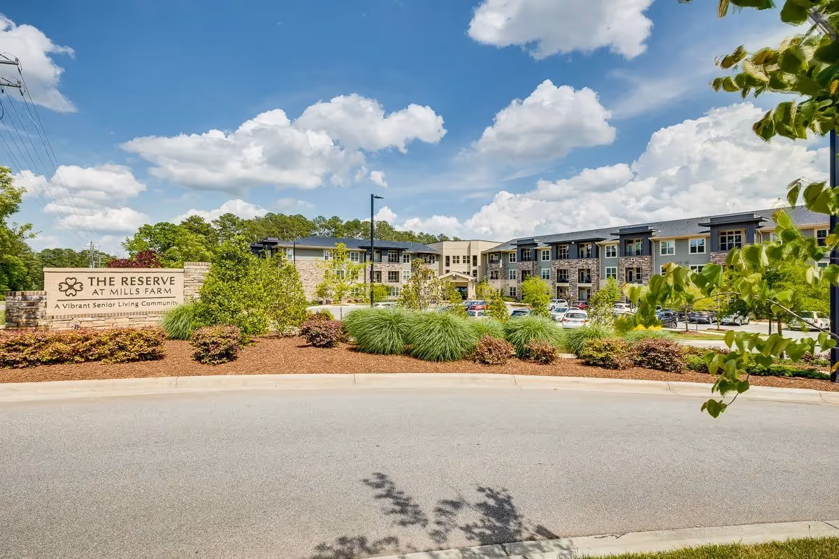 Exterior view of The Reserve at Mills Farm senior living community building with a clear blue sky and scattered clouds. The building is surrounded by landscaped greenery, bushes, and trees, with a parking lot visible in front.