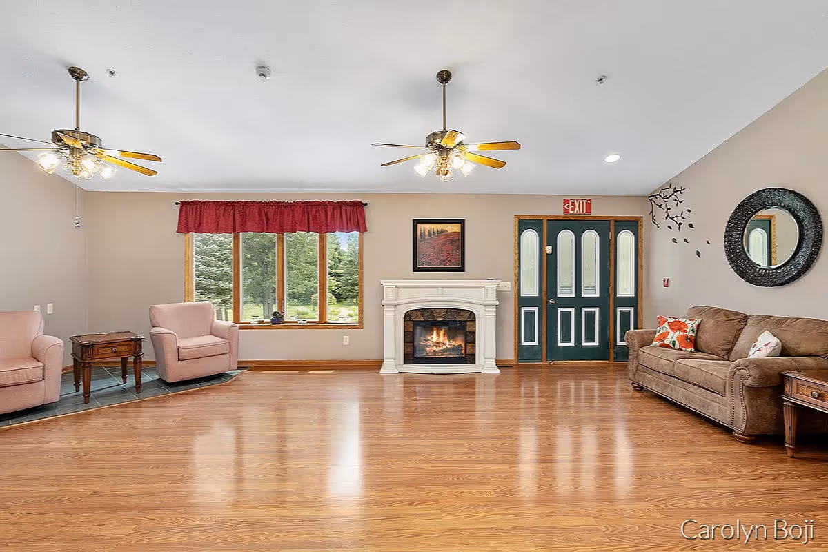 A spacious living room with wooden flooring, two ceiling fans with lights, a white fireplace with a fire burning, a window with a red valance showing greenery outside, two pink armchairs with a small wooden side table between them, a brown sofa with colorful pillows, a round black-framed mirror on the wall, and a green double door with an exit sign above it.