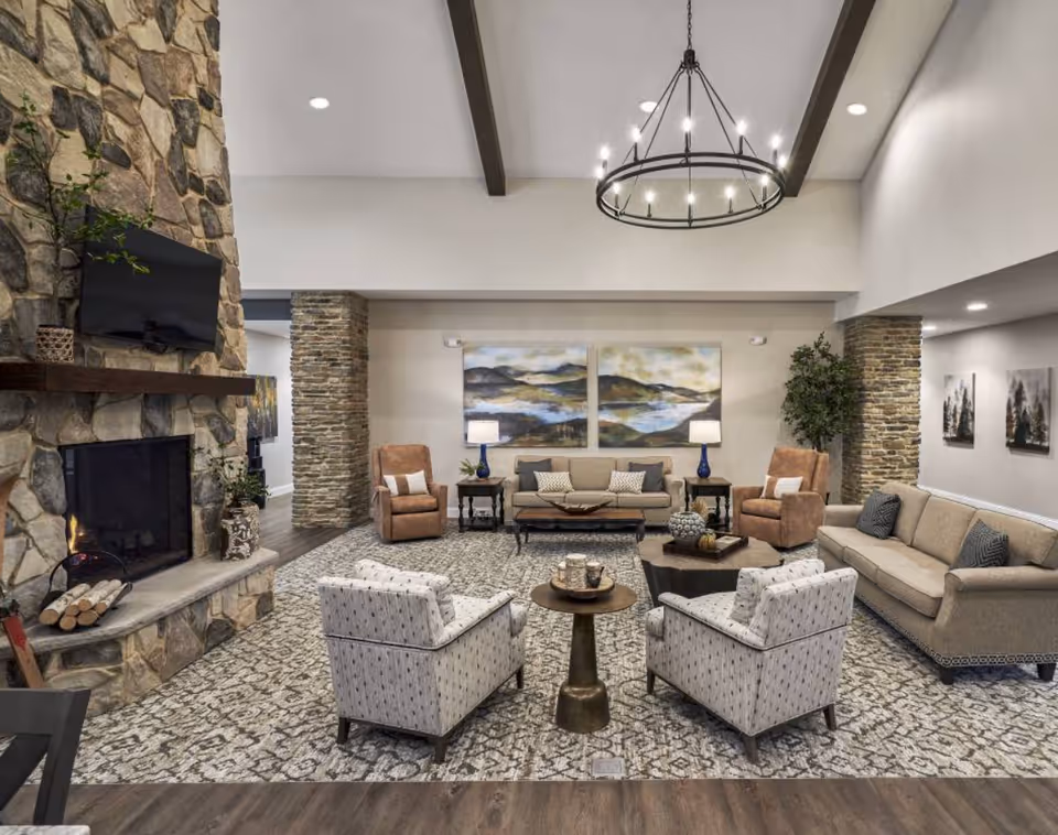A spacious senior living facility common area with a stone fireplace on the left, a mounted TV above it, and a cozy fire burning. The room features a patterned carpet, two beige sofas, two patterned armchairs, two brown leather recliners, and several side tables with lamps and decorative items. The walls have landscape paintings and there is a large circular chandelier hanging from the ceiling with exposed beams.