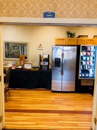 View of a small bistro area with a stainless steel refrigerator, a vending machine stocked with snacks, a coffee machine on a black-covered table, and wooden flooring. The area is framed by a doorway with a sign above that reads 'BISTRO'.
