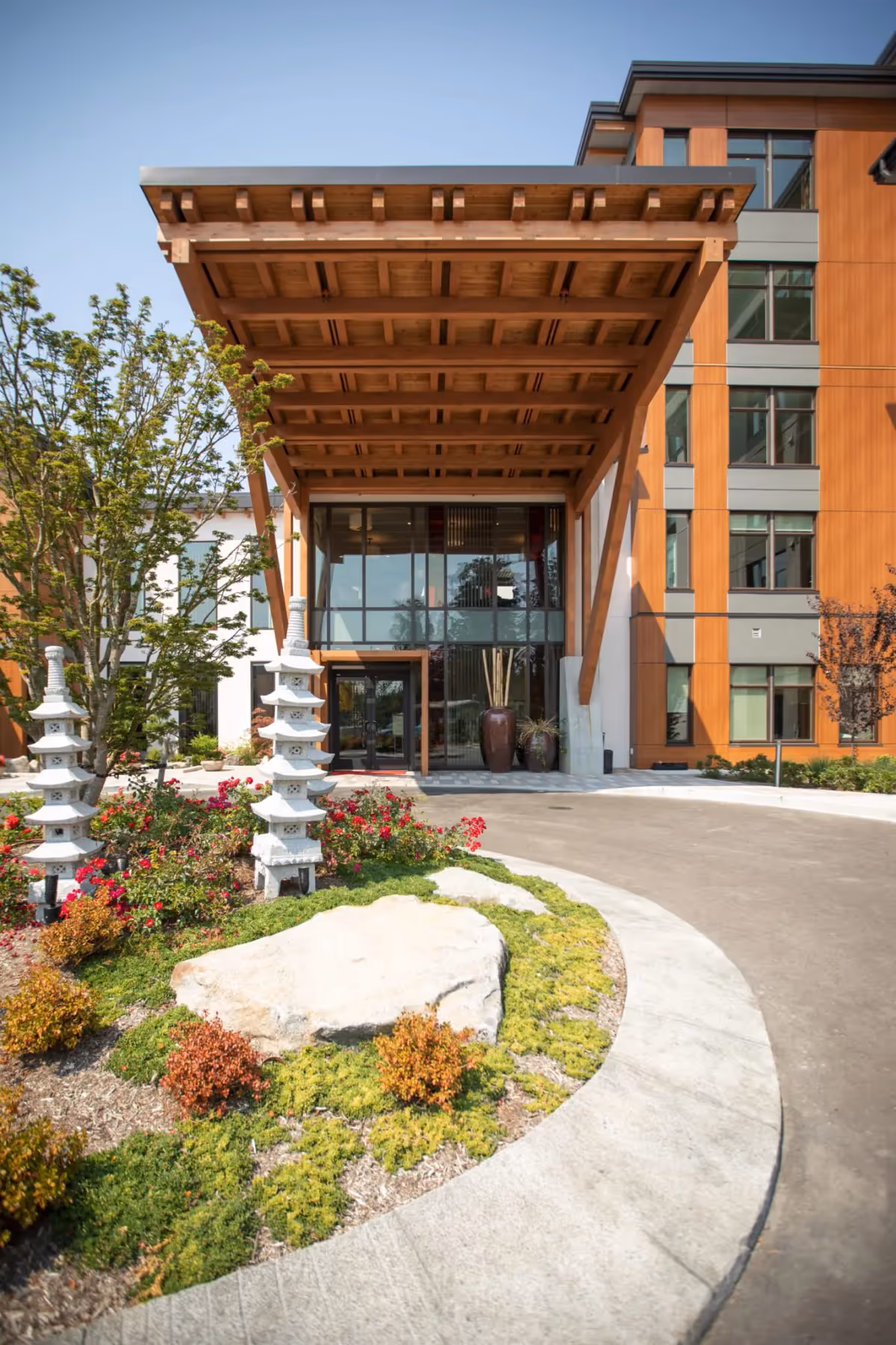 Entrance of Aegis Gardens Newcastle facility featuring a large wooden canopy supported by angled beams, glass doors, and windows. In front of the entrance is a landscaped circular garden with rocks, shrubs, red flowers, and two white stone pagoda-style sculptures.