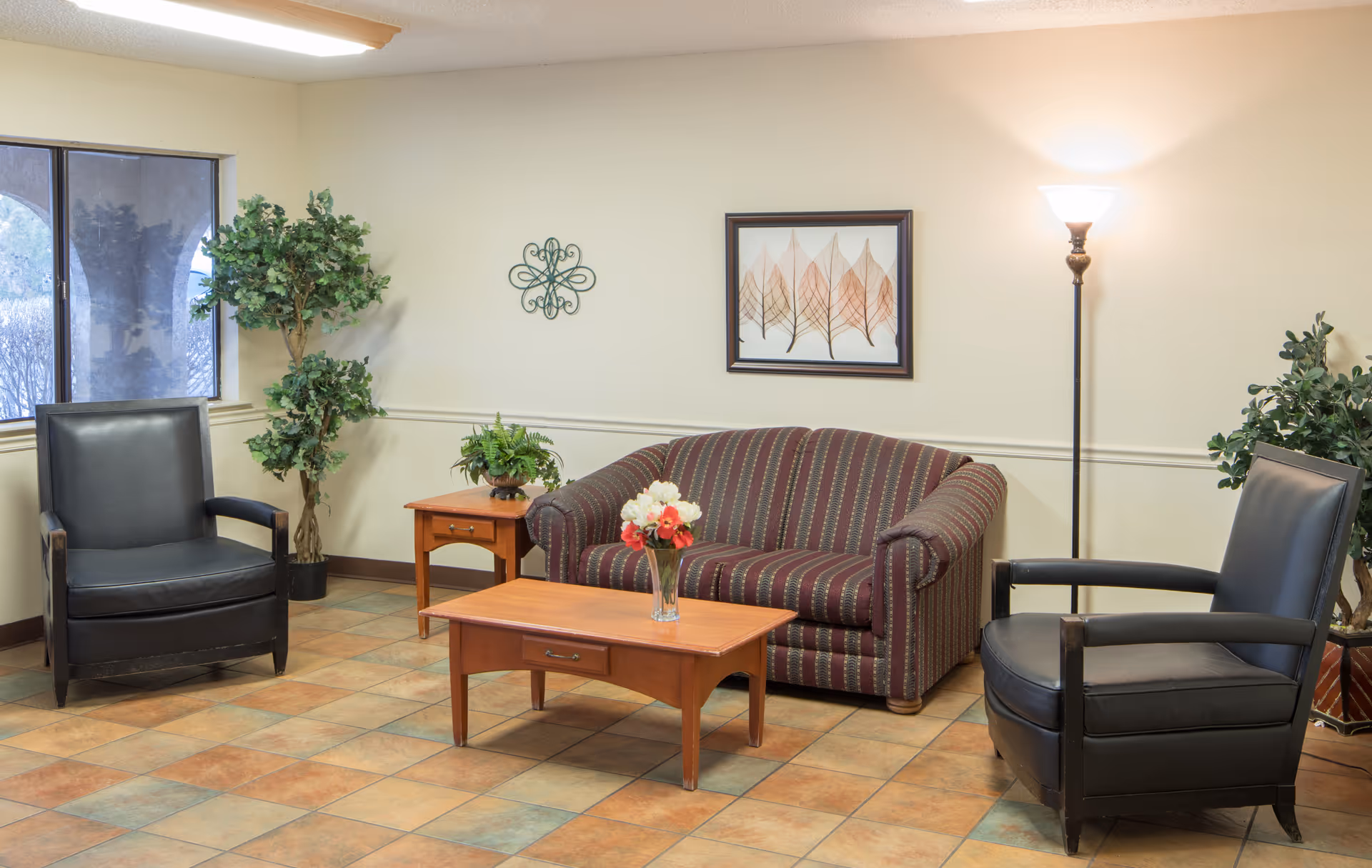 A cozy sitting area with a striped loveseat, two black leather armchairs, a wooden coffee table with a vase of flowers, a wooden side table with a potted plant, a floor lamp, wall art featuring leaves, and two large potted plants near a window.