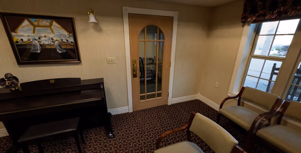 A small room with patterned carpet, beige walls, and a wooden door with glass panels. There is a piano with a bench on the left side, a painting above the piano, and three upholstered chairs with wooden armrests near a window on the right side. Outside the window, a rocking chair and part of a porch are visible.