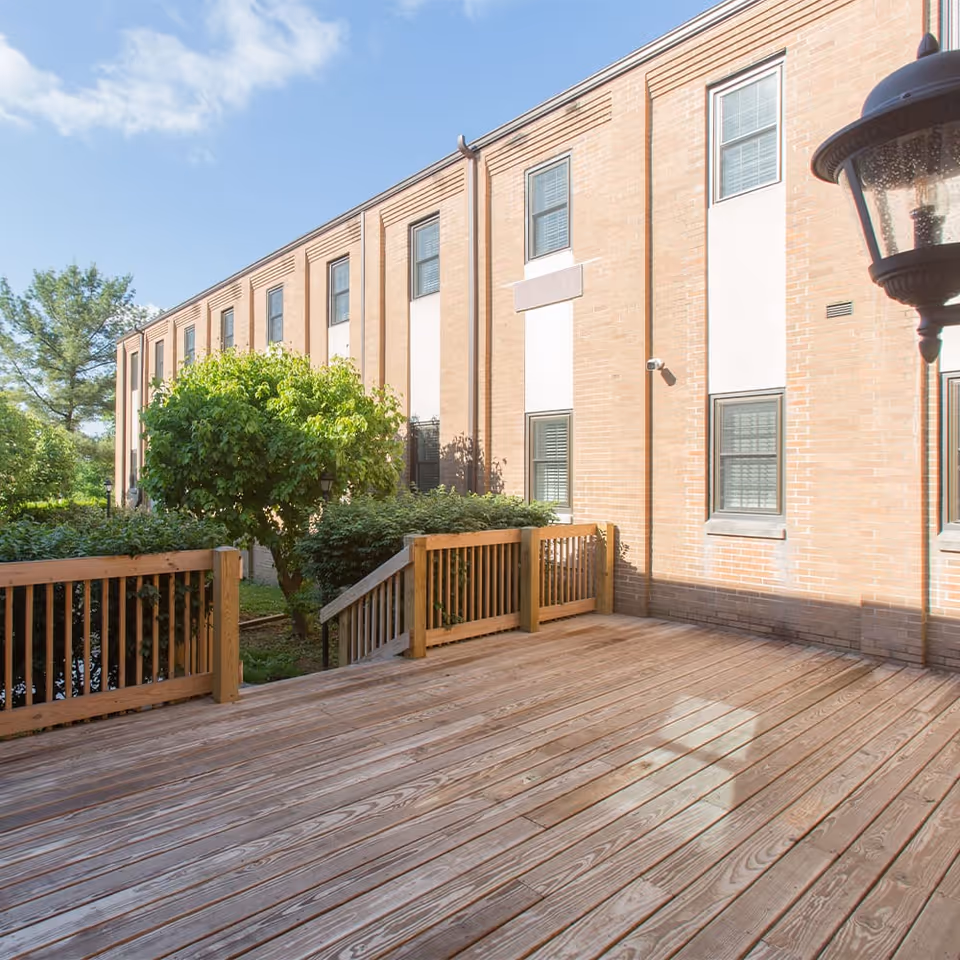 Outdoor wooden deck area with railing adjacent to a brick building with multiple windows, surrounded by green bushes and trees under a partly cloudy sky.