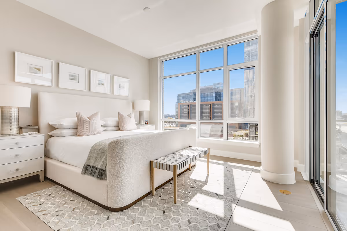 Bright modern bedroom with an upholstered bed, bedside tables, a bench, and large floor-to-ceiling windows overlooking the city.