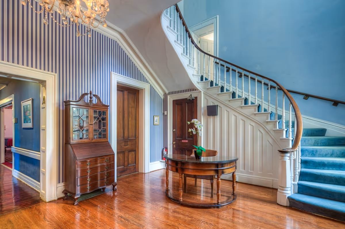 Interior view of a senior living facility featuring a curved staircase with blue carpeting and white railing. The walls are painted blue with white paneling under the stairs and blue and white vertical striped wallpaper on the adjacent wall. There is a wooden secretary desk with glass doors and a round wooden table with a small potted plant on it. The floor is polished hardwood, and a chandelier is partially visible at the top left corner.