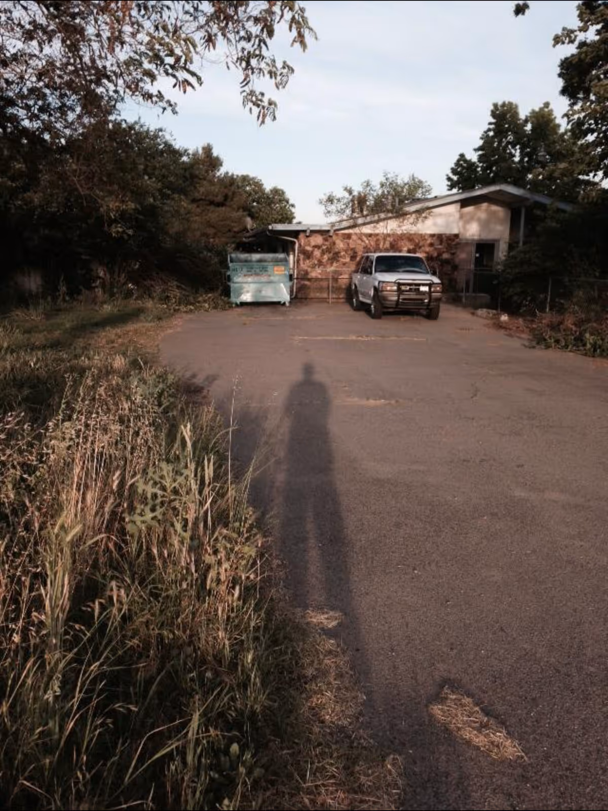 Parking lot in front of a low single-story building with a pickup truck, a dumpster, and a long shadow on the pavement.