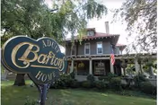 Exterior view of Barton's Adult Home, a large two-story house with a covered porch, surrounded by trees and greenery. An American flag is displayed on the porch, and a decorative sign with the facility's name is prominently placed on the lawn in front.