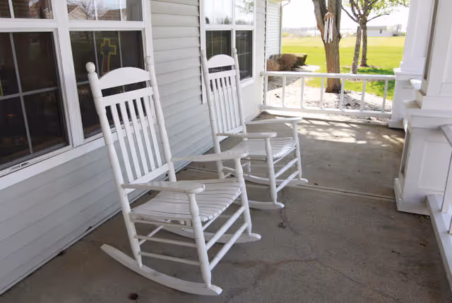 Two white wooden rocking chairs on a covered porch outside a building with windows, overlooking a grassy area with trees in the background.