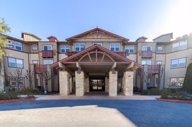 Front exterior view of a three-story senior living facility building with a covered entrance supported by stone pillars, multiple windows, and small balconies. The sky is clear and blue.
