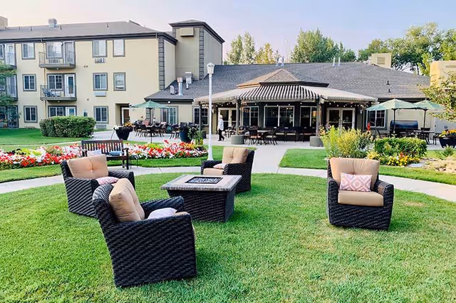 Outdoor seating area on a green lawn with four cushioned wicker chairs arranged around a square fire pit table. In the background, there is a building with a covered patio area featuring tables and chairs, umbrellas, and flower beds with colorful flowers.
