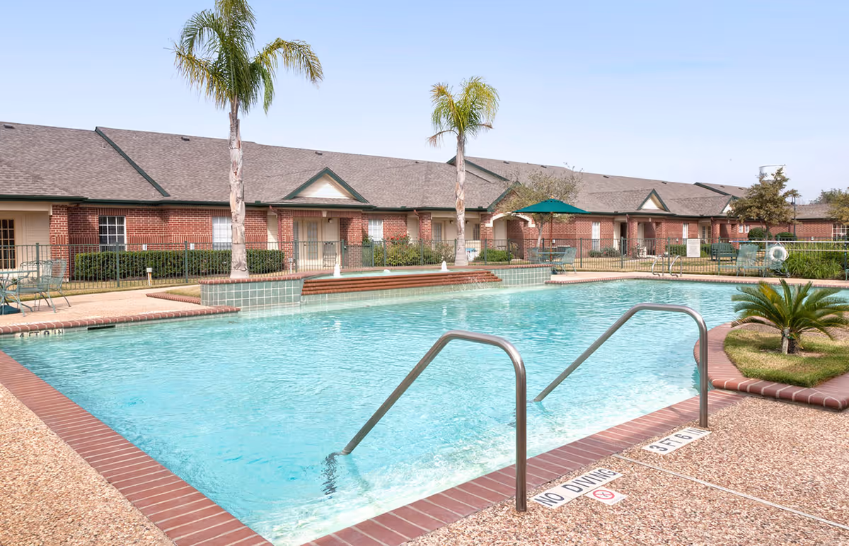 Outdoor swimming pool with clear blue water surrounded by a textured concrete deck and brick edging. Two metal handrails lead into the pool. In the background, there are single-story brick buildings with green trim, palm trees, patio tables with umbrellas, and some greenery under a clear blue sky.