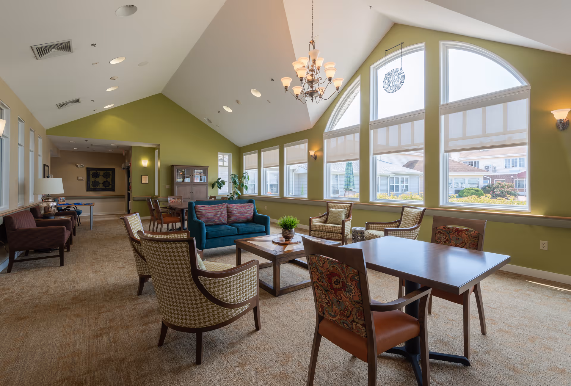 Sunlit communal living room with a vaulted ceiling and large arched windows, furnished with sofas, armchairs, tables, and a coffee table.
