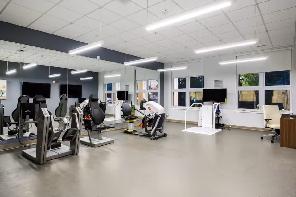 A spacious fitness room with exercise machines including seated resistance equipment, a large mirror wall, multiple windows letting in natural light, two wall-mounted TVs, and a desk with an office chair in the corner.
