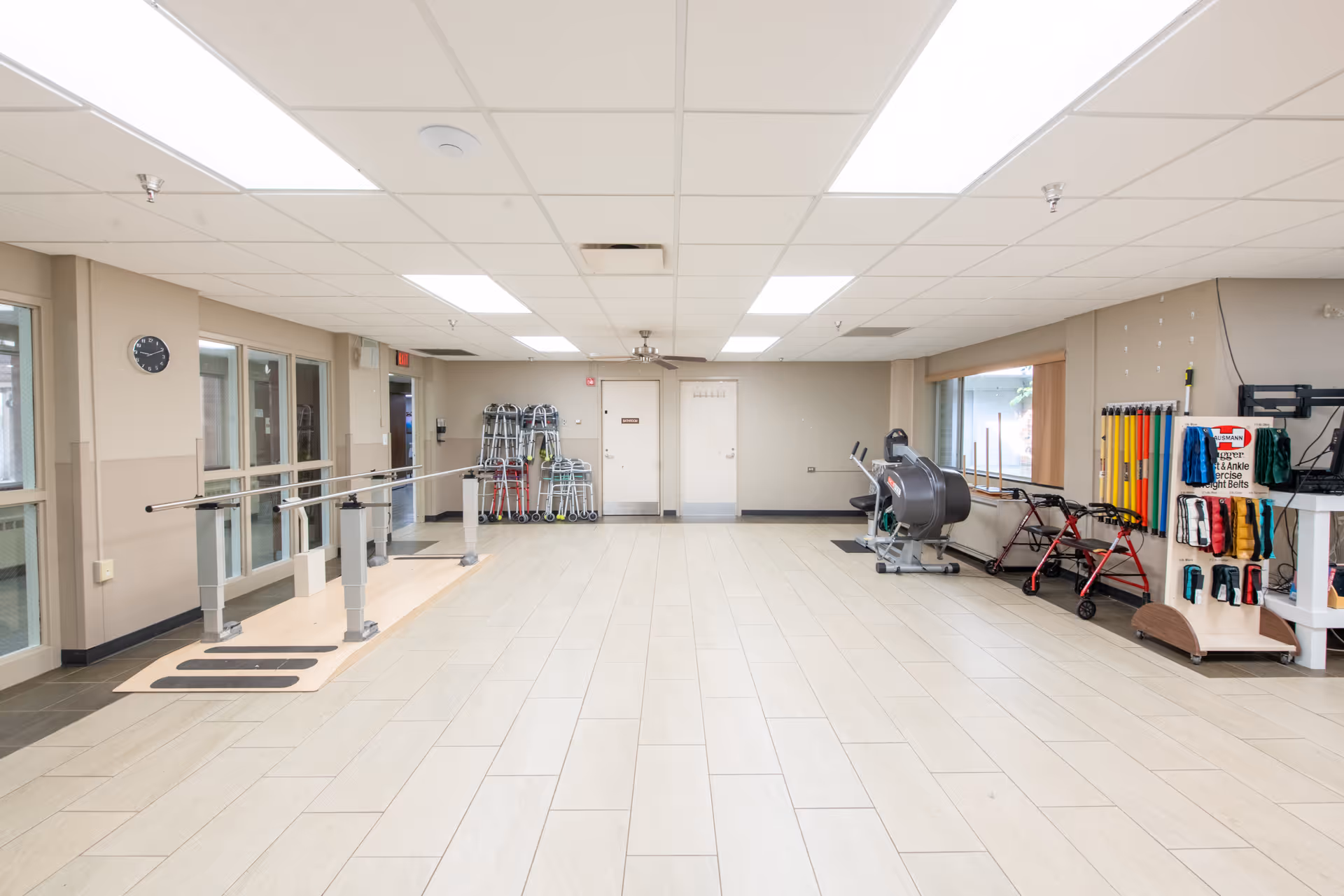 Bright rehabilitation/exercise room with parallel bars, walkers, rollators, exercise machines and therapy supplies.