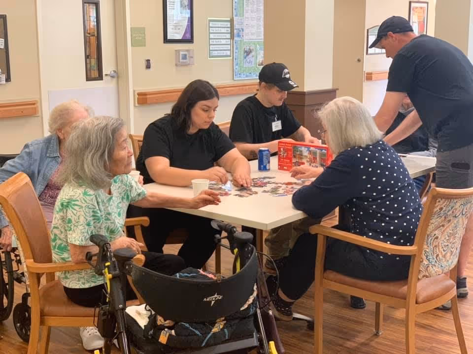 A group of elderly individuals and two younger adults sitting around a table working on a jigsaw puzzle in a well-lit room with wooden floors and beige walls. One elderly woman is seated in a wheelchair, and another person is standing nearby. The atmosphere appears to be calm and social.