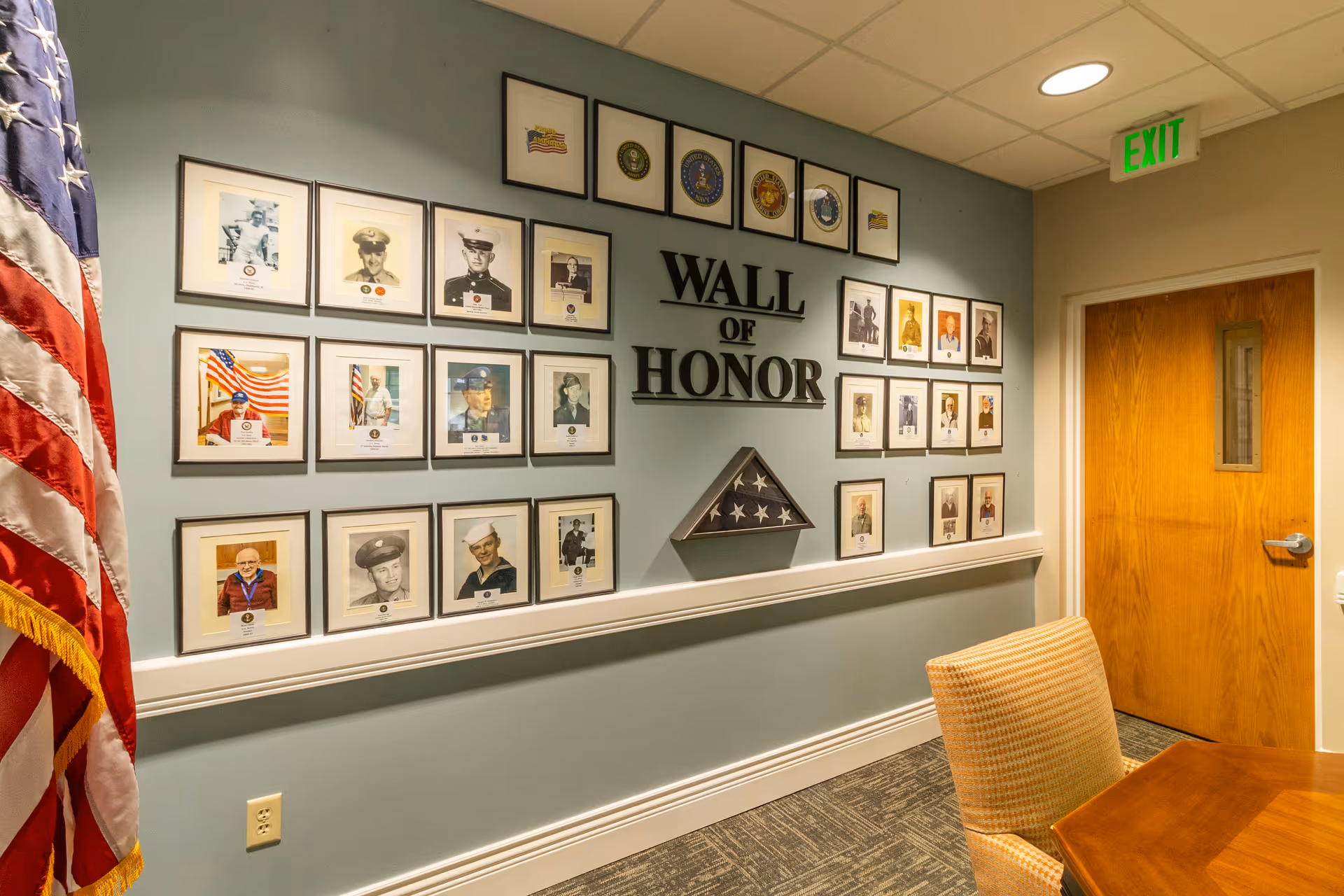 Interior hallway with a Wall of Honor display featuring framed photographs of veterans and military insignias on a light blue wall. An American flag is partially visible on the left side, and there is a wooden door with a small window and an illuminated green exit sign above it on the right. A patterned chair and a dark table are in the foreground.