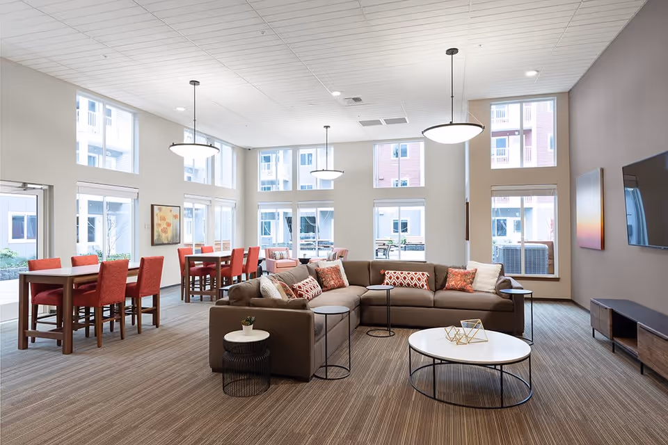 A spacious and modern living room area with a large brown sectional sofa adorned with patterned throw pillows, a round white coffee table with decorative items, and several small side tables. The room features high ceilings with hanging pendant lights, large windows letting in natural light, and multiple dining tables with red chairs in the background. A flat-screen TV is mounted on the wall opposite the sofa.