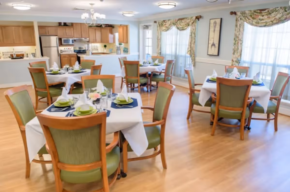 A bright dining room with several round tables covered with white tablecloths, each set with green plates, napkins, and glassware. The room has wooden chairs with green cushions, large windows with floral curtains, and a kitchen area with wooden cabinets and stainless steel appliances in the background.