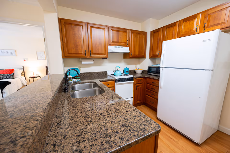 Small kitchen with wooden cabinets, granite countertops, a double sink, white refrigerator and stove, with a bedroom visible through an open doorway.