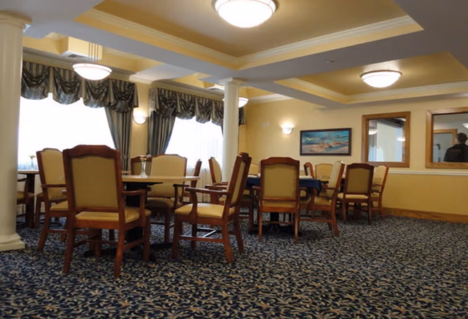 A dining room with several wooden tables and cushioned chairs arranged on a patterned carpet. The room has yellow walls, large windows with draped curtains, ceiling lights, and framed artwork on the wall.
