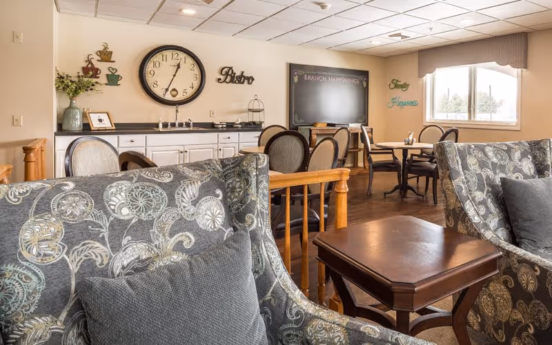A cozy common area in a senior living facility featuring patterned armchairs with gray cushions, a wooden side table, and a dining area with round tables and chairs. The wall has a large clock, decorative coffee cup wall art, and the word 'Bistro'. A chalkboard labeled 'Branch Happenings' and a window letting in natural light are also visible.