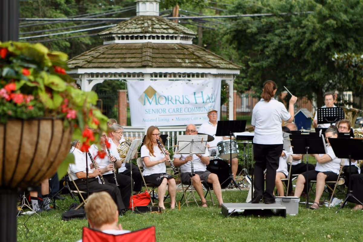 A group of musicians performing outdoors in front of a white gazebo with a banner that reads 'Morris Hall Senior Care Community'. The musicians are seated on chairs on a grassy area, playing various wind and percussion instruments, while a conductor stands facing them. There are trees and bushes in the background and a hanging flower basket in the foreground.