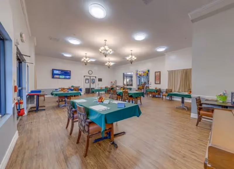 A spacious dining room with multiple tables covered in green tablecloths, each set with placemats and small centerpieces. The room has wooden flooring, several chandeliers and ceiling lights, and large windows on the left side. There is a wall-mounted TV and double doors at the far end of the room.