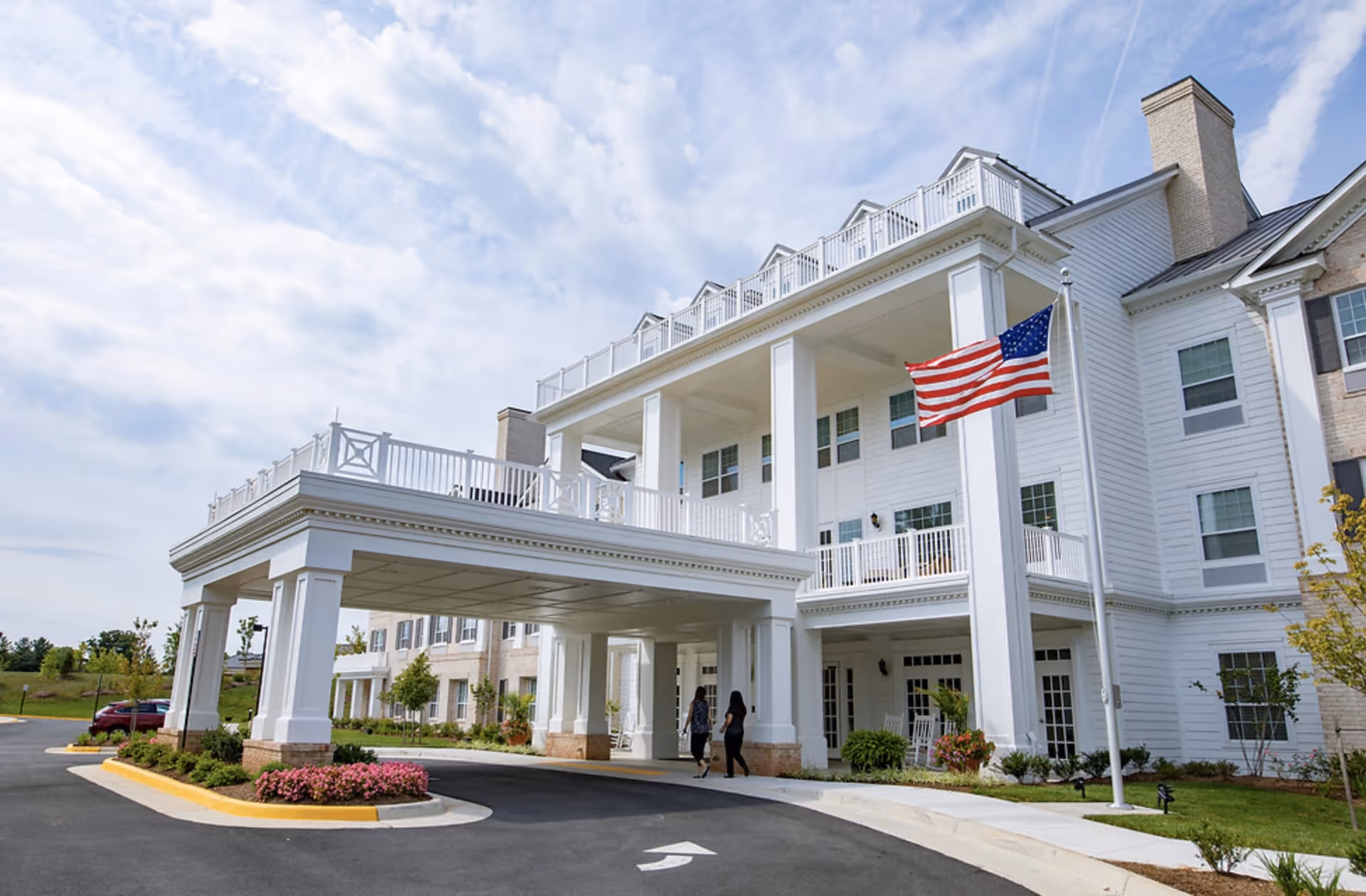 The front exterior of The Wellington at Lake Manassas senior living facility showing a large white building with multiple windows, a covered entrance supported by columns, an American flag on a flagpole, and two people walking near the entrance under a partly cloudy sky.