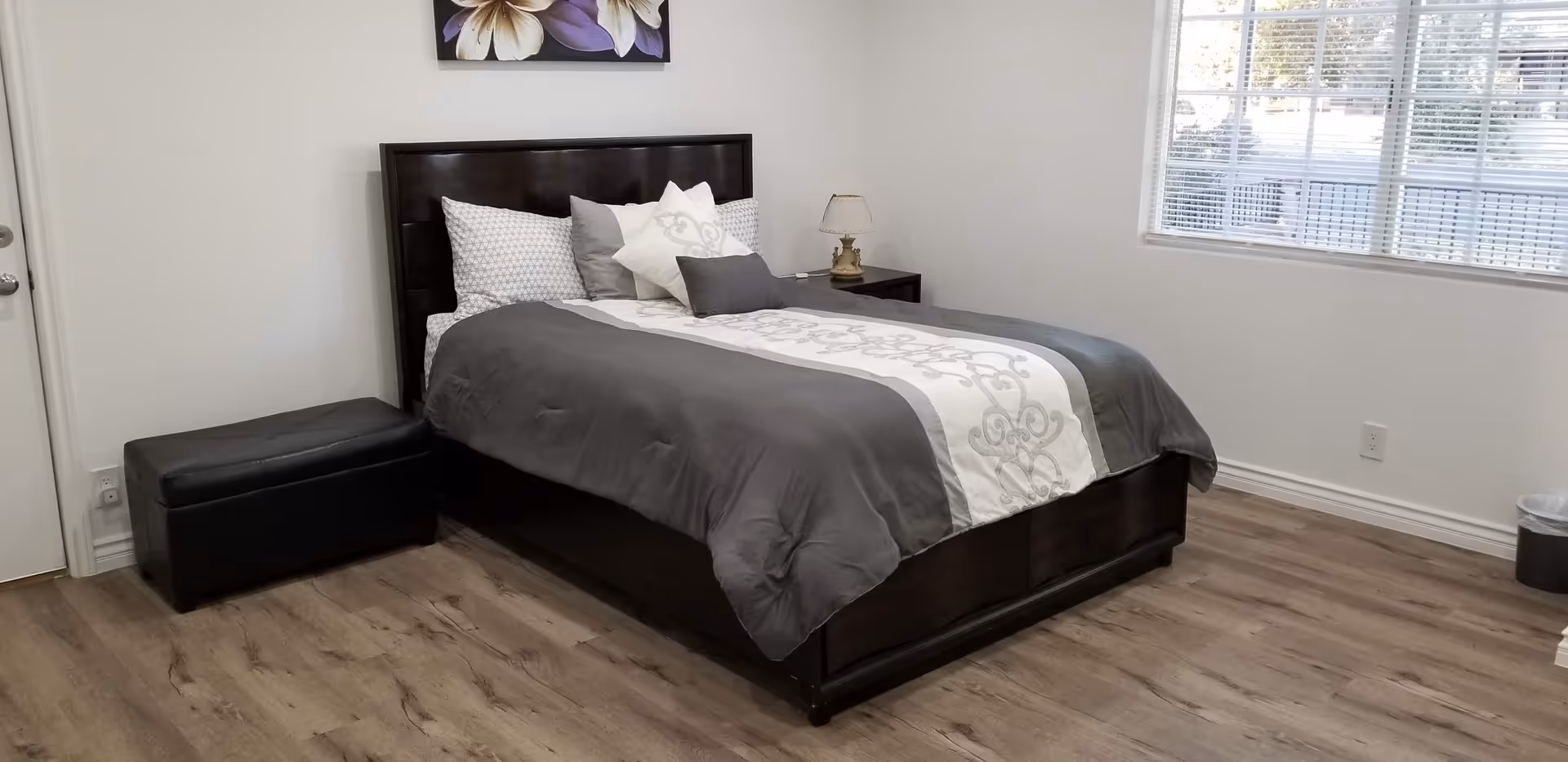 A neatly made bed with gray and white bedding in a bedroom with wooden flooring, a window with blinds, a small bedside table with a lamp, a black storage ottoman, and a floral painting on the wall.