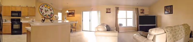 A spacious assisted living common area featuring a kitchen with wooden cabinets, black appliances, and a countertop island. Adjacent to the kitchen is a dining area with a table and chairs. The living room area includes a beige leather sofa, a large flat-screen TV, and windows letting in natural light. The walls are decorated with framed artwork, and an exit door is visible in the background.