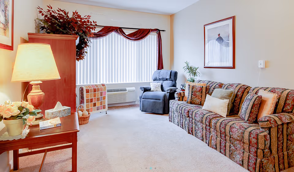 A cozy living room with a floral patterned sofa adorned with multiple cushions, a blue recliner chair, a wooden side table with a lamp, flowers, and books, a wooden cabinet with a potted plant on top, and a large window with vertical blinds and a red valance.