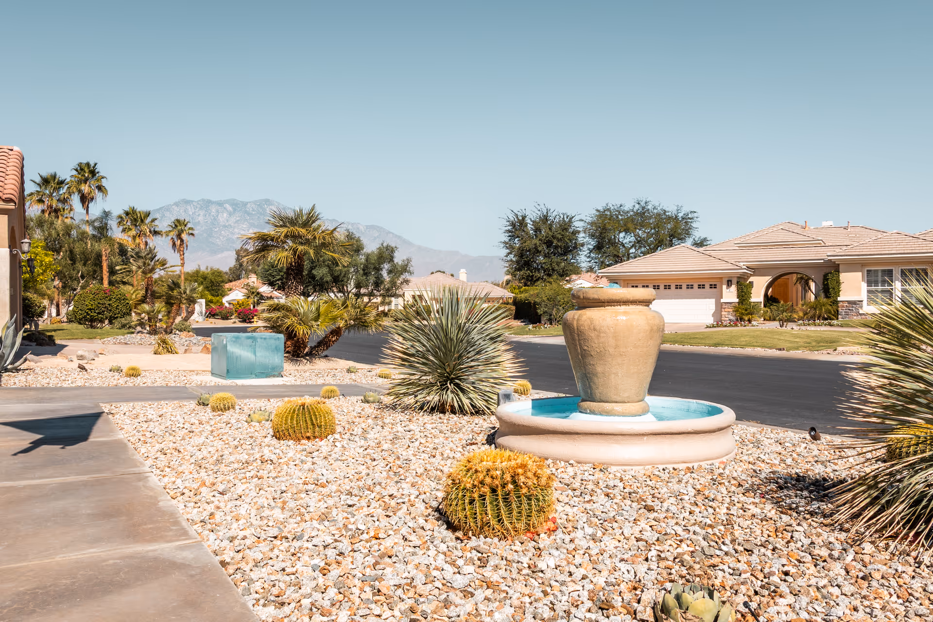 Desert landscaped street with a central stone fountain, cacti and palm trees in front of single-story homes with mountains in the background.