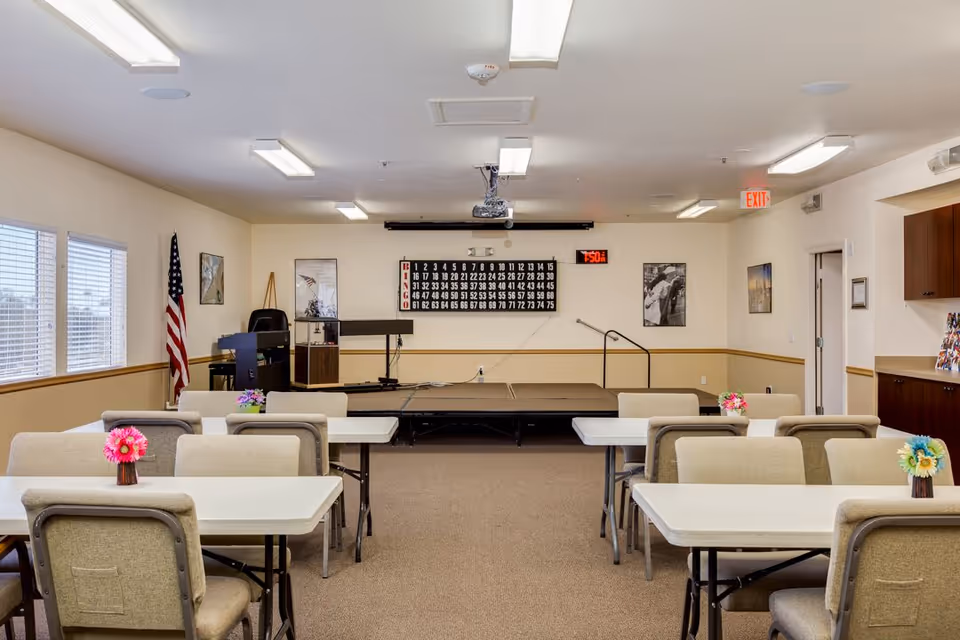 A community room with several tables and chairs arranged in rows facing a small stage. The stage has a bingo board and a digital clock displaying 7:50. There are framed pictures on the walls, an American flag in the corner, and small flower arrangements on each table. The room is well-lit with ceiling lights and has windows on the left side.