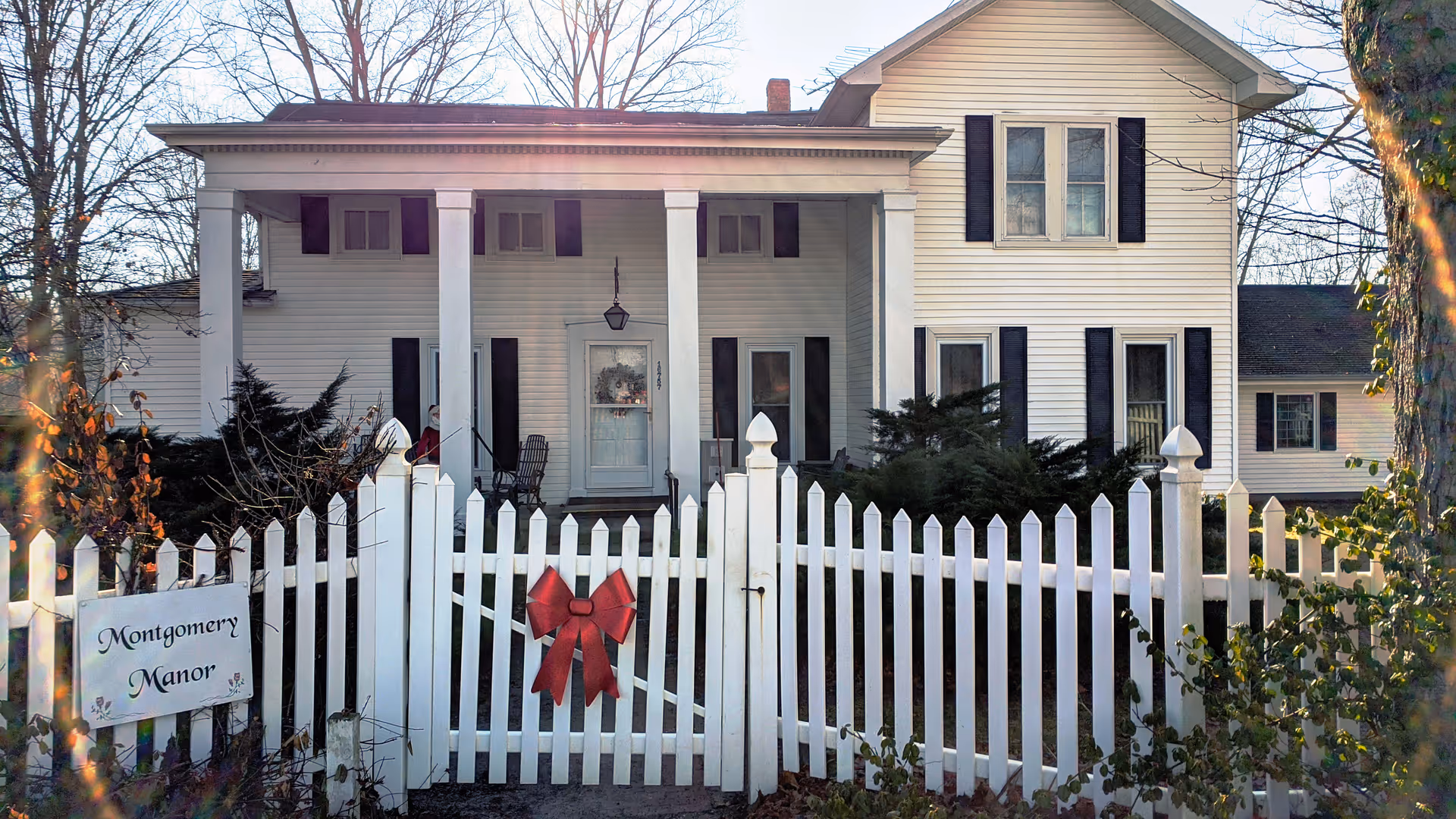 Front exterior view of Montgomery Manor, a white two-story house with black shutters, a porch with columns, and a white picket fence decorated with a red bow.
