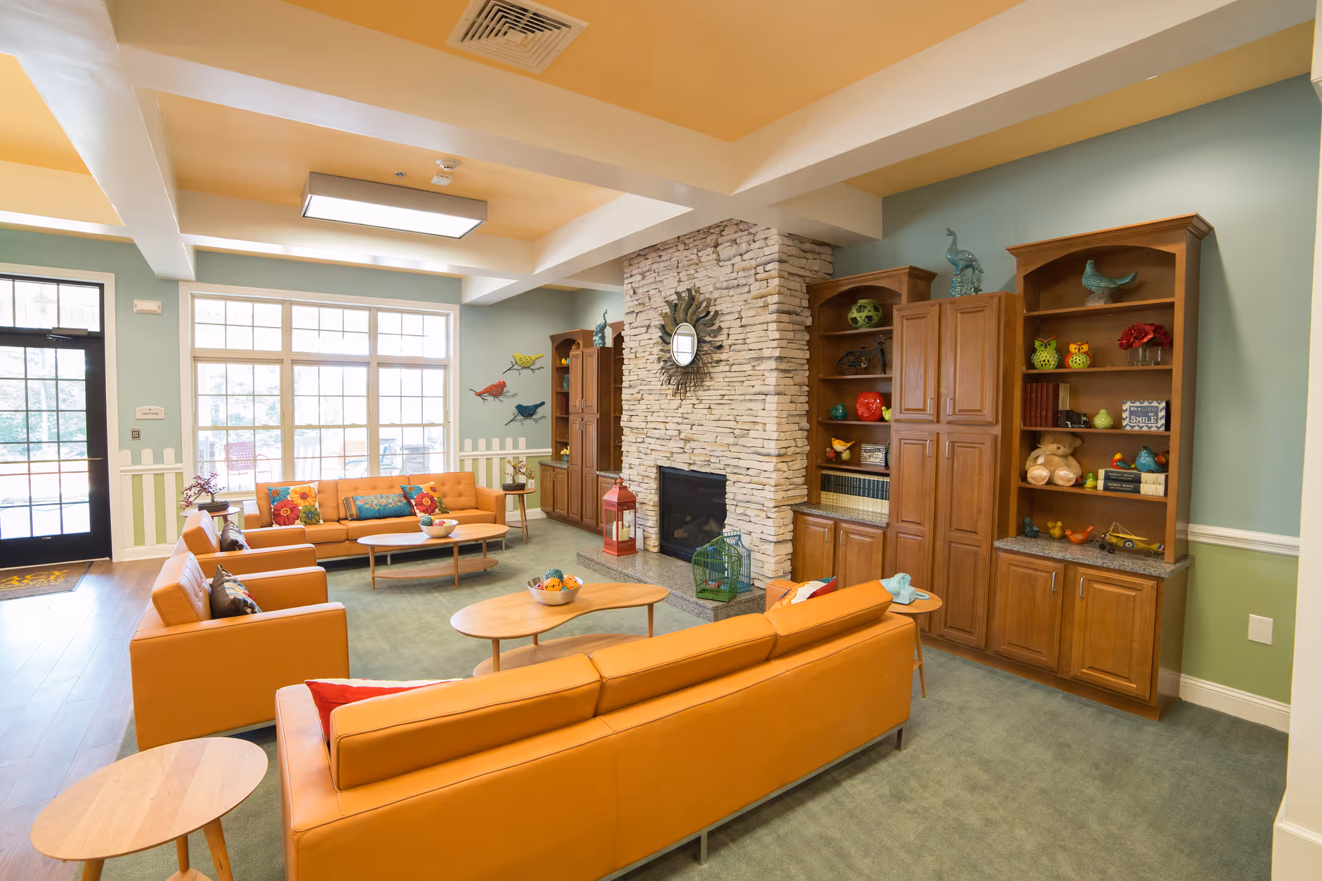 A bright and spacious living room with large windows letting in natural light. The room features orange leather sofas and armchairs arranged around wooden coffee tables. A stone fireplace with a decorative sunburst mirror above it is centered between two wooden cabinets filled with colorful decorative items and books. The walls are painted in soft green and blue tones, and the floor is carpeted in a muted green color.