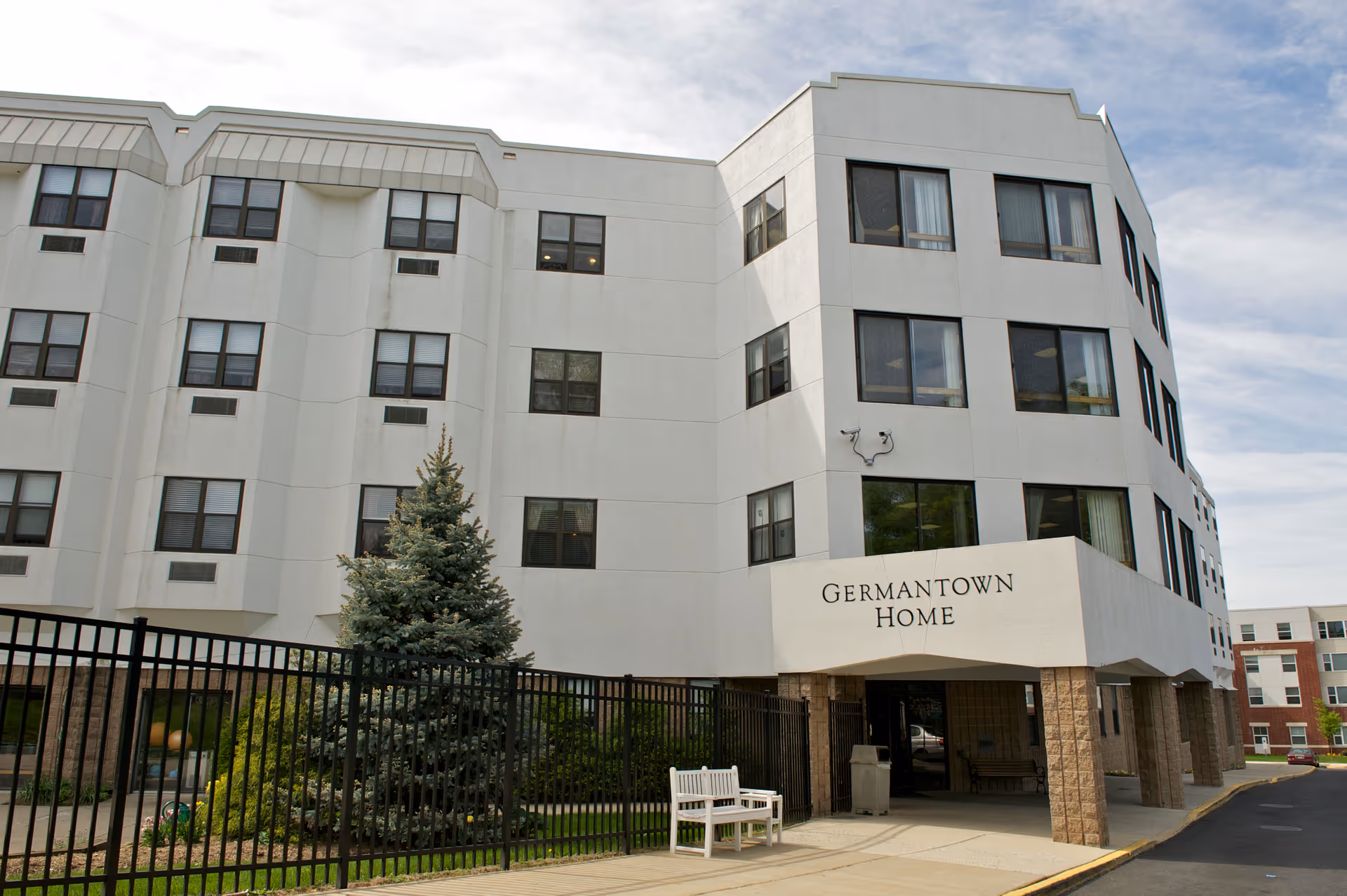 White multi-story senior living building with an entrance canopy reading "Germantown Home", fenced landscaping and a bench outside.
