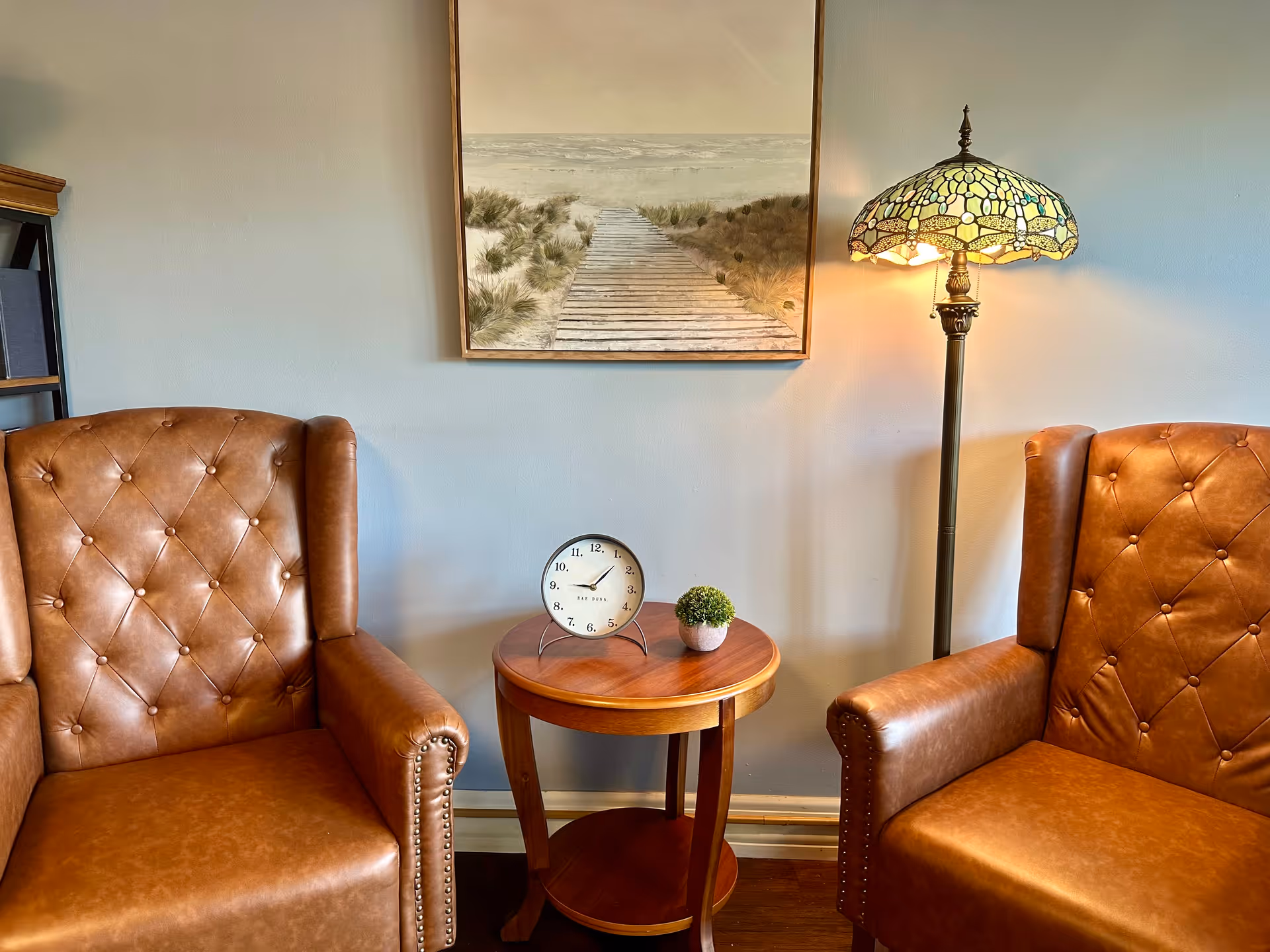 Two brown leather armchairs with button tufting are positioned on either side of a small round wooden table. On the table, there is a small round clock and a small potted plant. A Tiffany-style floor lamp with a stained glass shade is on the right side, and a framed painting of a wooden boardwalk leading through sand dunes to the ocean hangs on the wall behind.