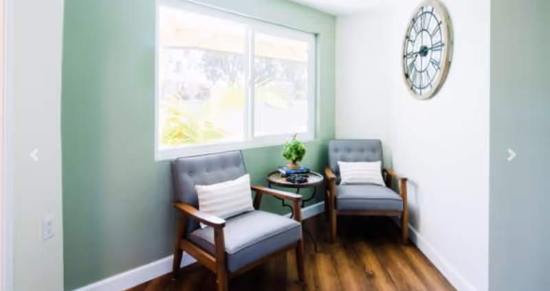 A bright sitting area with two gray armchairs, a small round side table by a window, and a large wall clock.