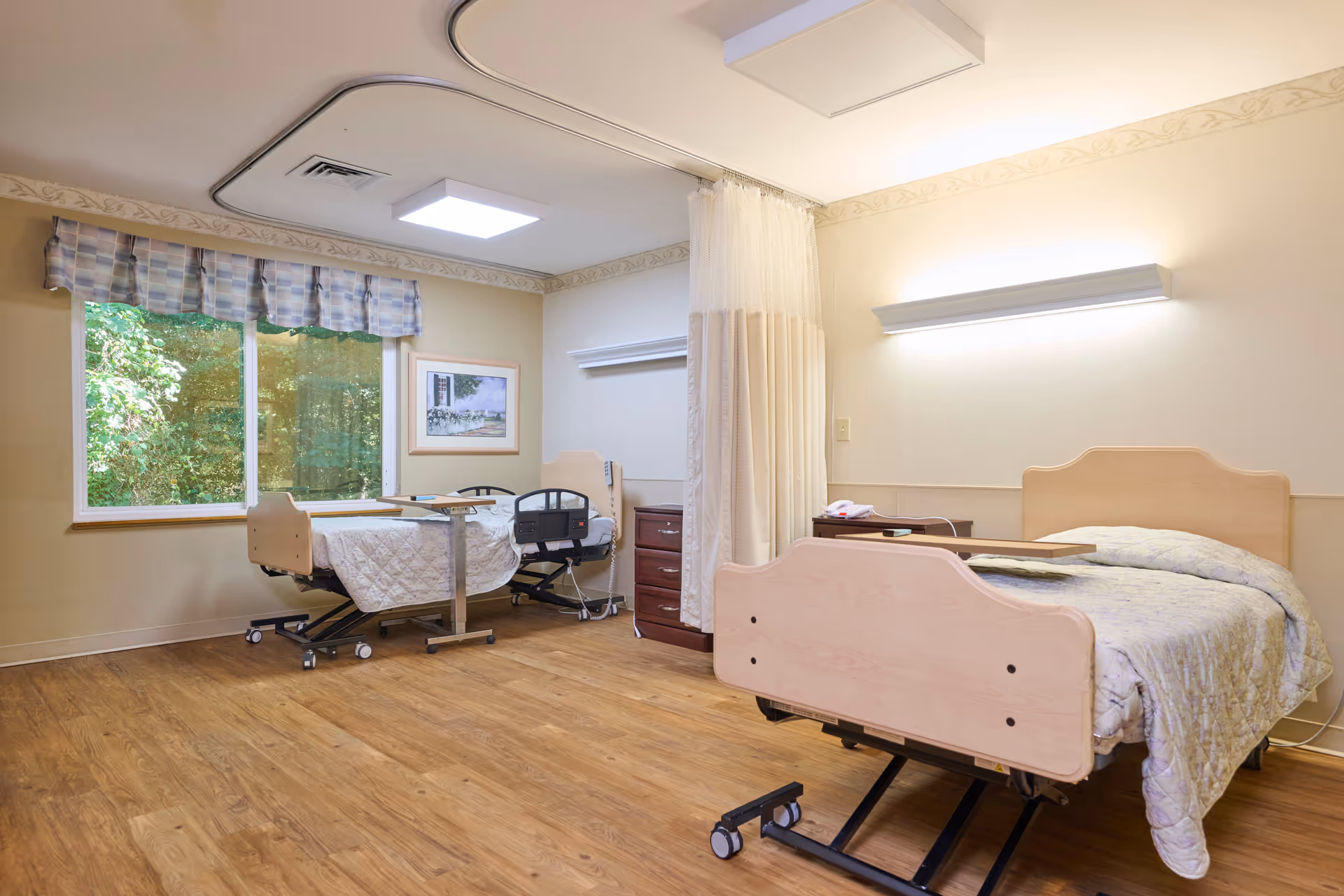 A nursing facility room with two hospital beds, each with a light-colored wooden headboard and footboard. One bed is positioned near a large window with a plaid valance, showing greenery outside. There is a privacy curtain between the beds, a wooden nightstand with a telephone, and overhead lighting on the ceiling and wall. The floor is wood-patterned, and the room has a calm, neutral color scheme.
