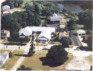 Aerial view of a single-story facility and surrounding grounds with trees, driveways, and nearby water.