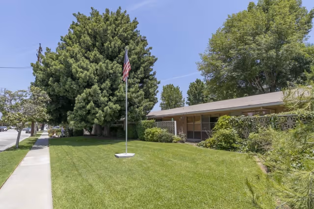 A single-story building with large windows partially covered by greenery, surrounded by a well-maintained lawn with a flagpole displaying the American flag in the center. There are large trees behind and around the building, and a sidewalk runs along the left side of the lawn under a clear blue sky.