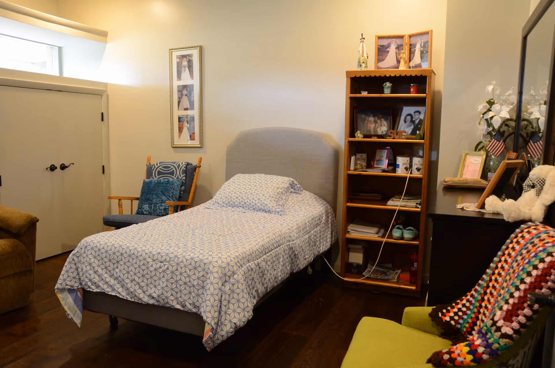 Single-occupancy bedroom with a neatly made twin bed, wooden bookshelf, chairs, and framed photos.