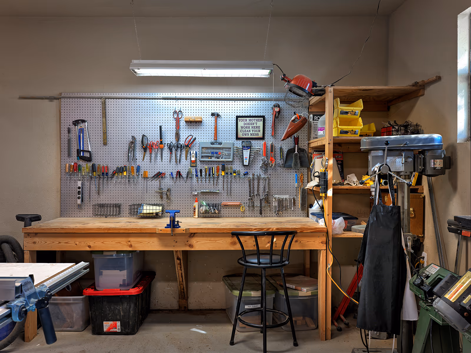 A tidy workshop with a wooden workbench, pegboard of tools, shelving and a stool under a fluorescent light.